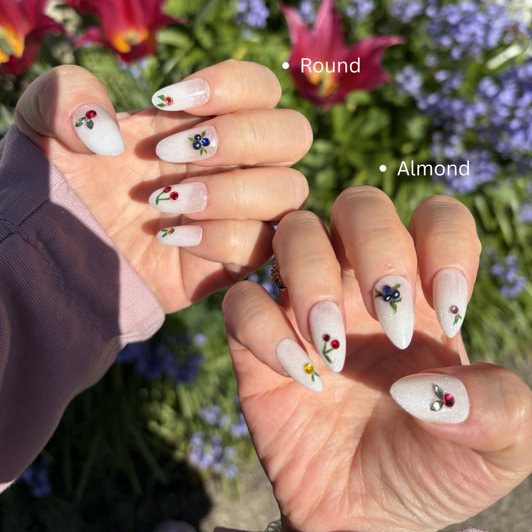 Close-up of hands with floral nail art . press-on nails feature delicate hand-painted cherries and blueberries, finished with brilliant authentic Swarovski charms on a shimmering white glitter base. Perfect for a dainty, premium, and summer-ready look.
