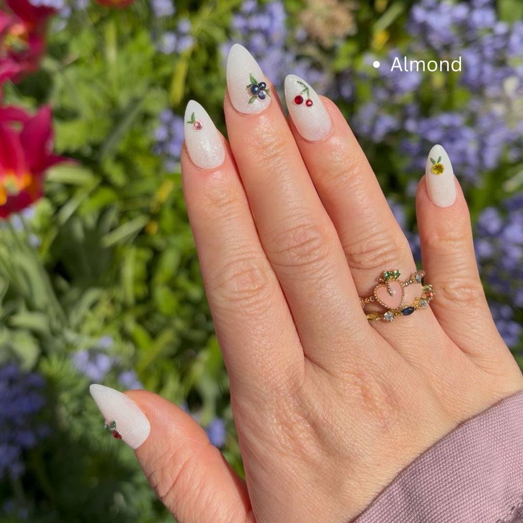 Hand with almond-shaped nails featuring decorative designs with cherry, blueberry and peach shaped Swarovski charms