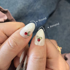 Close-up of hands with white nail polish and red gemstone decorations, showing round and almond nail shapes.