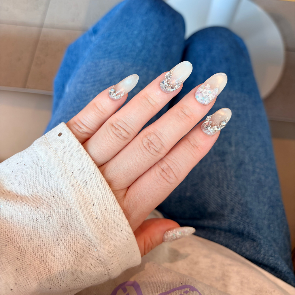 Hand with decorative nails showing off a clear nail polish with glittery accents.Reusable Swarovski nails, custom celestial press-ons, handmade luxury crystal nails, iridescent star gel nails.